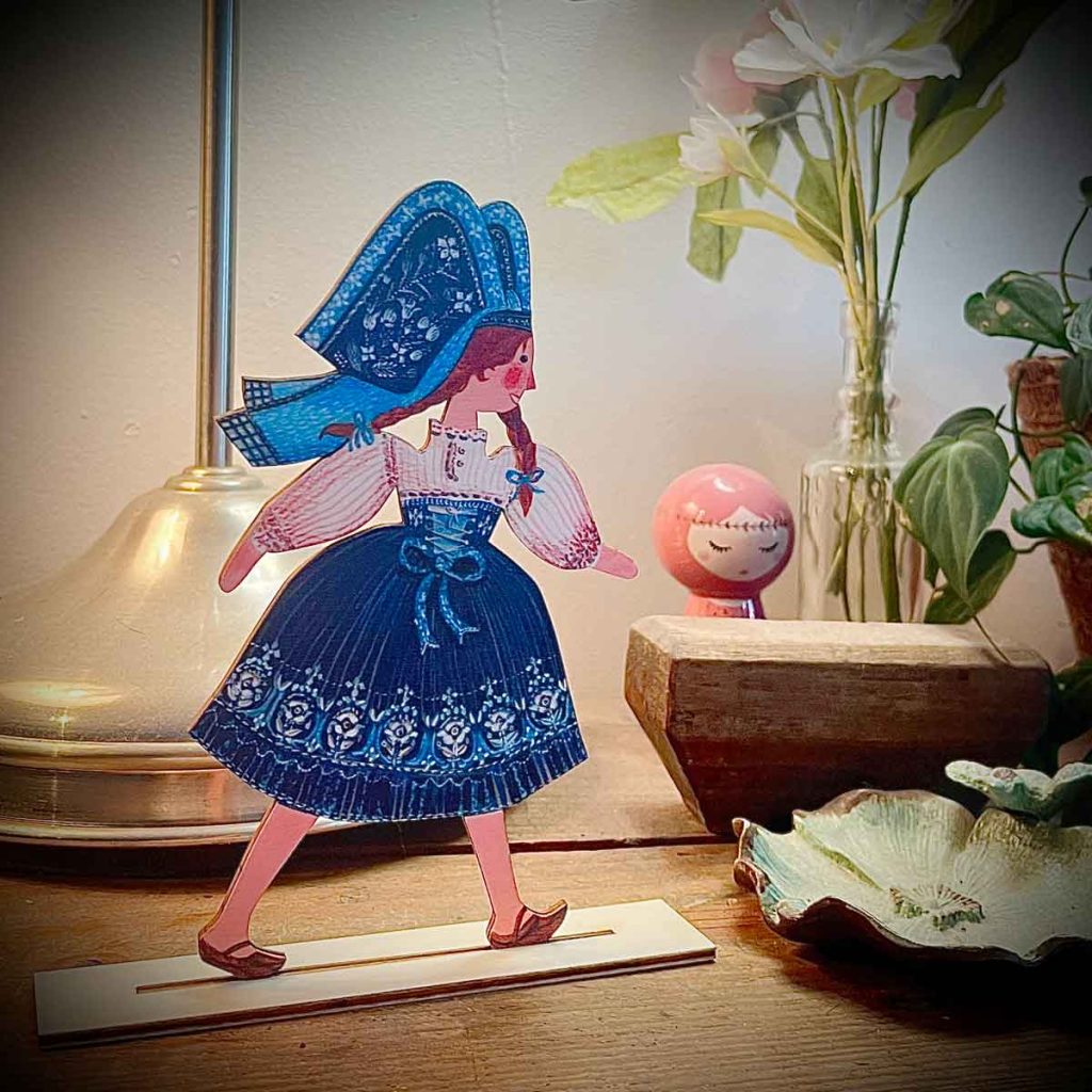 Figurine alsacienne illustrée en bois posée sur un meuble, entre une lampe, des fleurs et une petite poupée – Claire et Mélanie