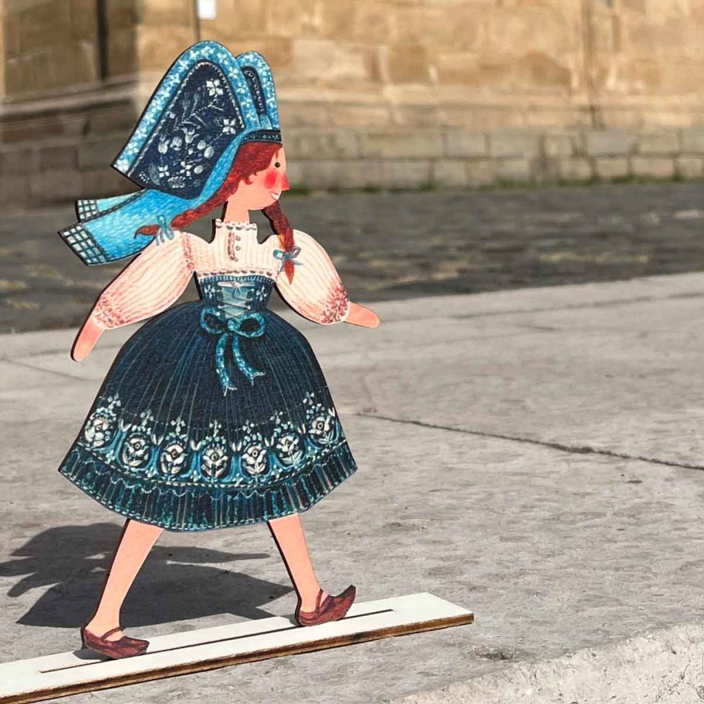 Suzel, figurine en bois d'une Alsacienne en costume traditionnel bleu sur son socle, en extérieur – Claire et Mélanie